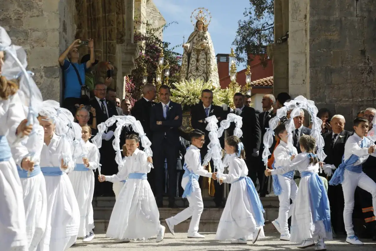 Ni&ntilde;os con trajes blancos y cinturones azules participan en una procesi&oacute;n, llevando arcos de encaje. Al fondo, la Virgen de la Gu&iacute;a es llevada en andas.