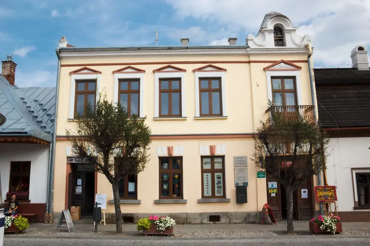 Stary Sącz rynek. Jasnoż&oacute;łta kamienica z brązowymi oknami i balkonem, przed nią drzewa i kwiaty.