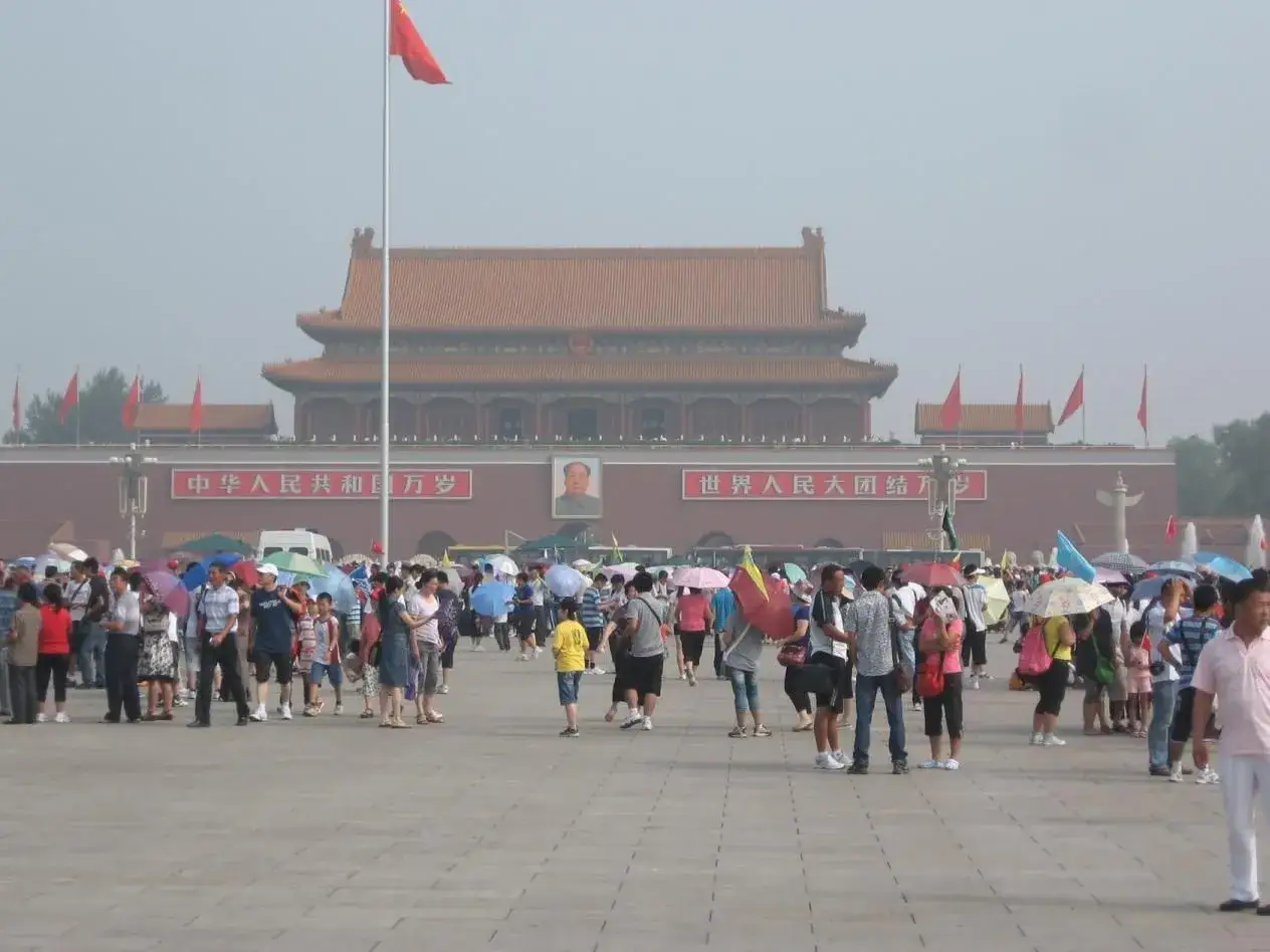 Tłum ludzi na placu **Tiananmen** pod czerwonym budynkiem z portretem Mao i chińskimi napisami.