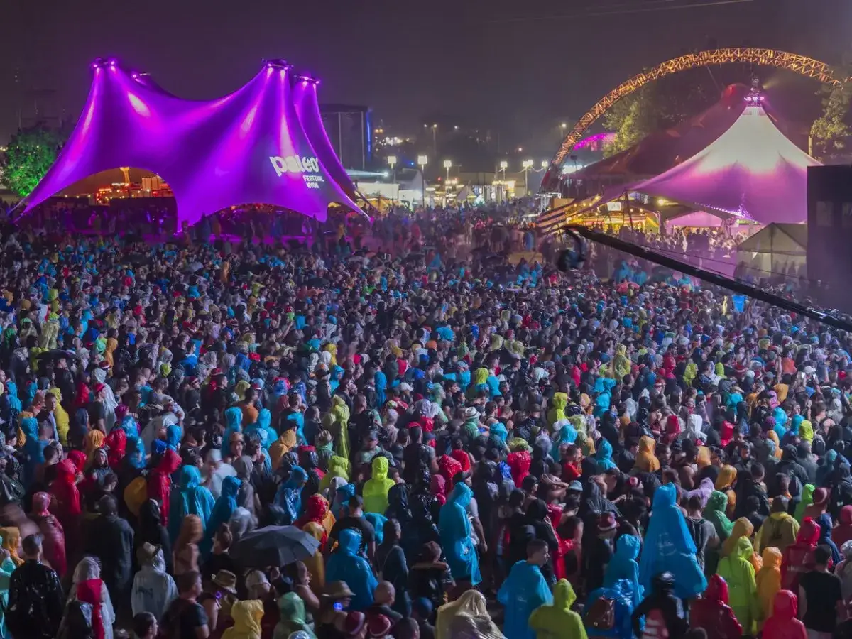 A massive crowd enjoys the vibrant atmosphere of the Greenfield Festival under a large, illuminated purple tent.