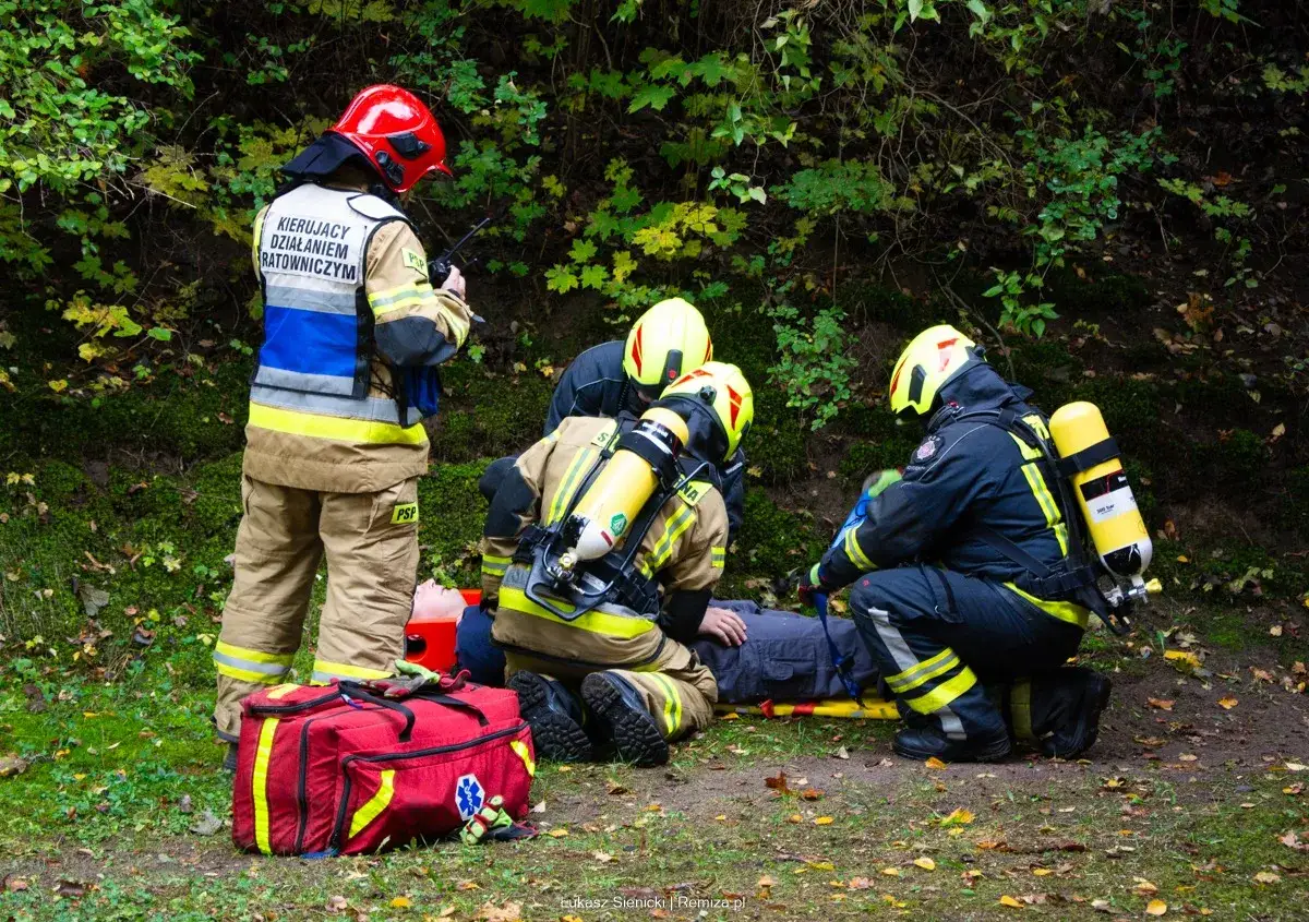 Dowódca sekcji OSP kieruje akcją ratunkową przy poszkodowanym w lesie. Strażacy udzielają pomocy.