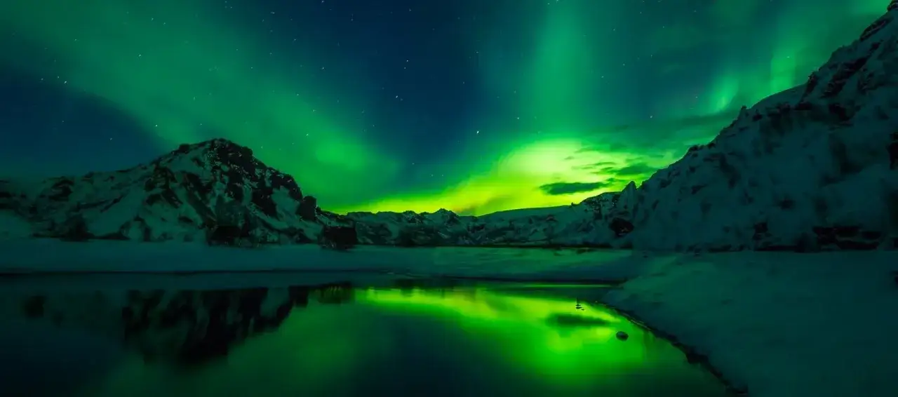 Islandia pogoda zimą: zorza polarna tańczy nad ośnieżonymi górami i spokojną wodą, odbijając zielone światła.