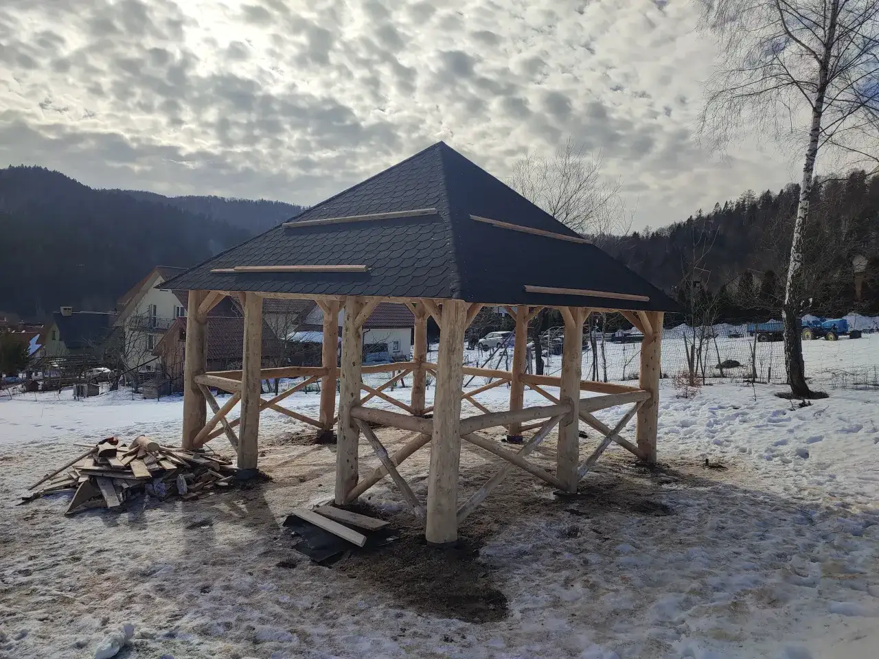 Drewniana altana w trakcie budowy, z ciemnym dachem z gontu. Wokół śnieg, stos desek. W tle góry i domy.