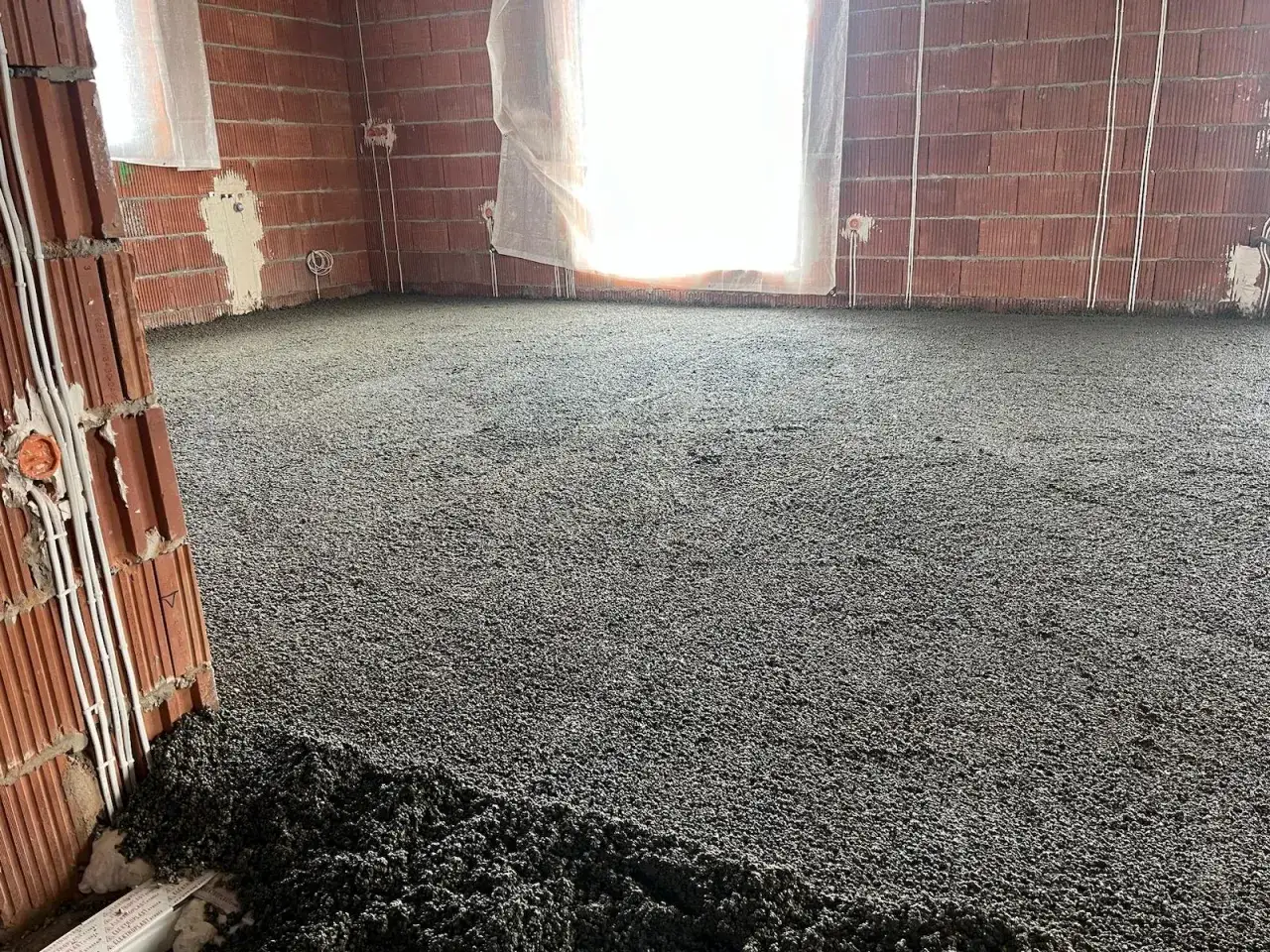 Świeżo wylany styrobeton na podłodze w budowanym pomieszczeniu. Widać ceglane ściany i okno.