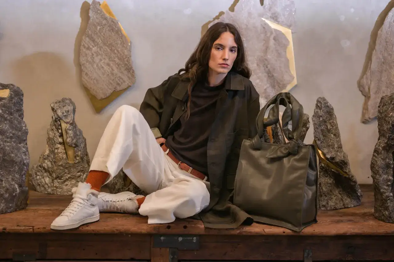 Eine Frau in l&auml;ssiger Kleidung, inspiriert von Officine Creative, sitzt vor skulpturalen Steinen. Sie tr&auml;gt eine olivgr&uuml;ne Jacke, wei&szlig;e Hose und Sneaker.