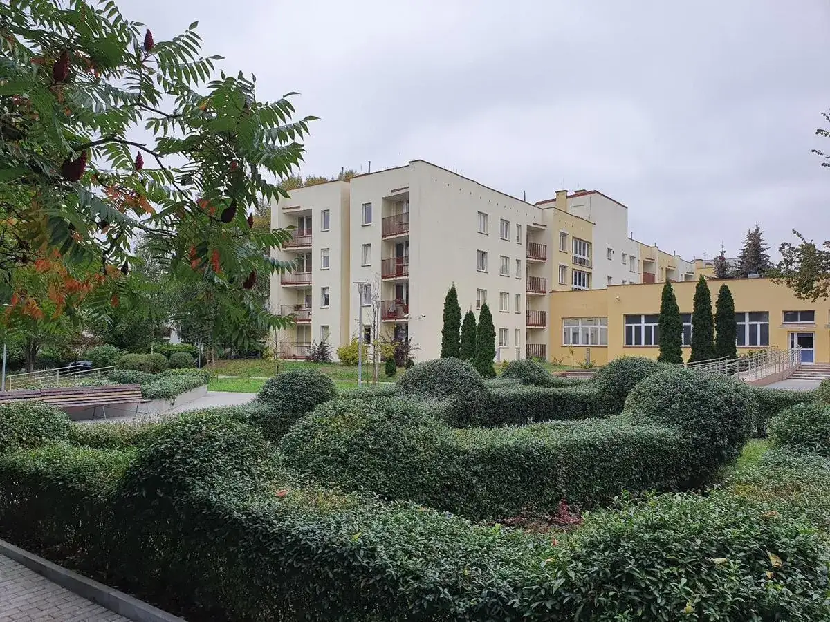 Zadbany teren zielony przed budynkiem, który może być domem emeryta wojskowego. Kręte żywopłoty i drzewa tworzą przyjazną przestrzeń.