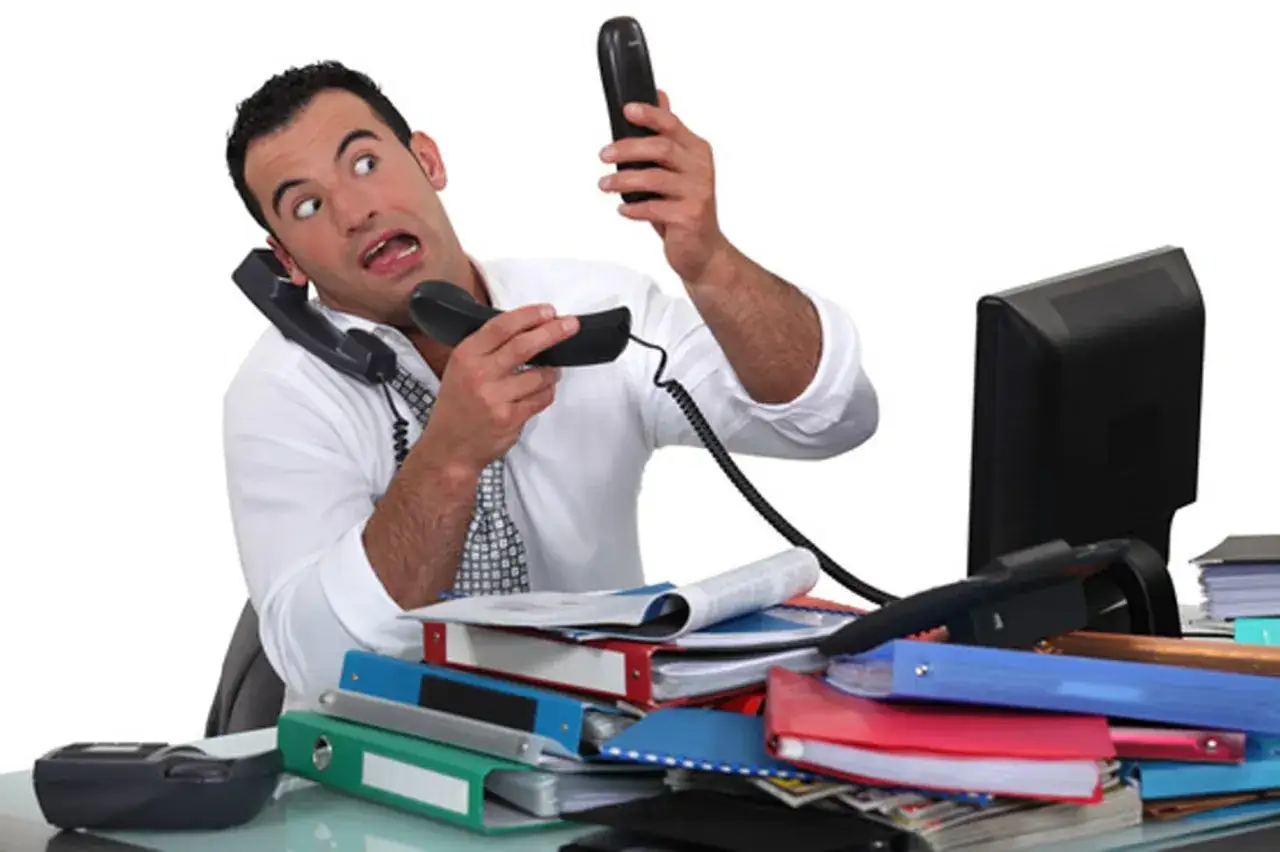 Uomo stressato con due telefoni in mano, cerca di rispondere al telefono in inglese mentre &egrave; sommerso da documenti.