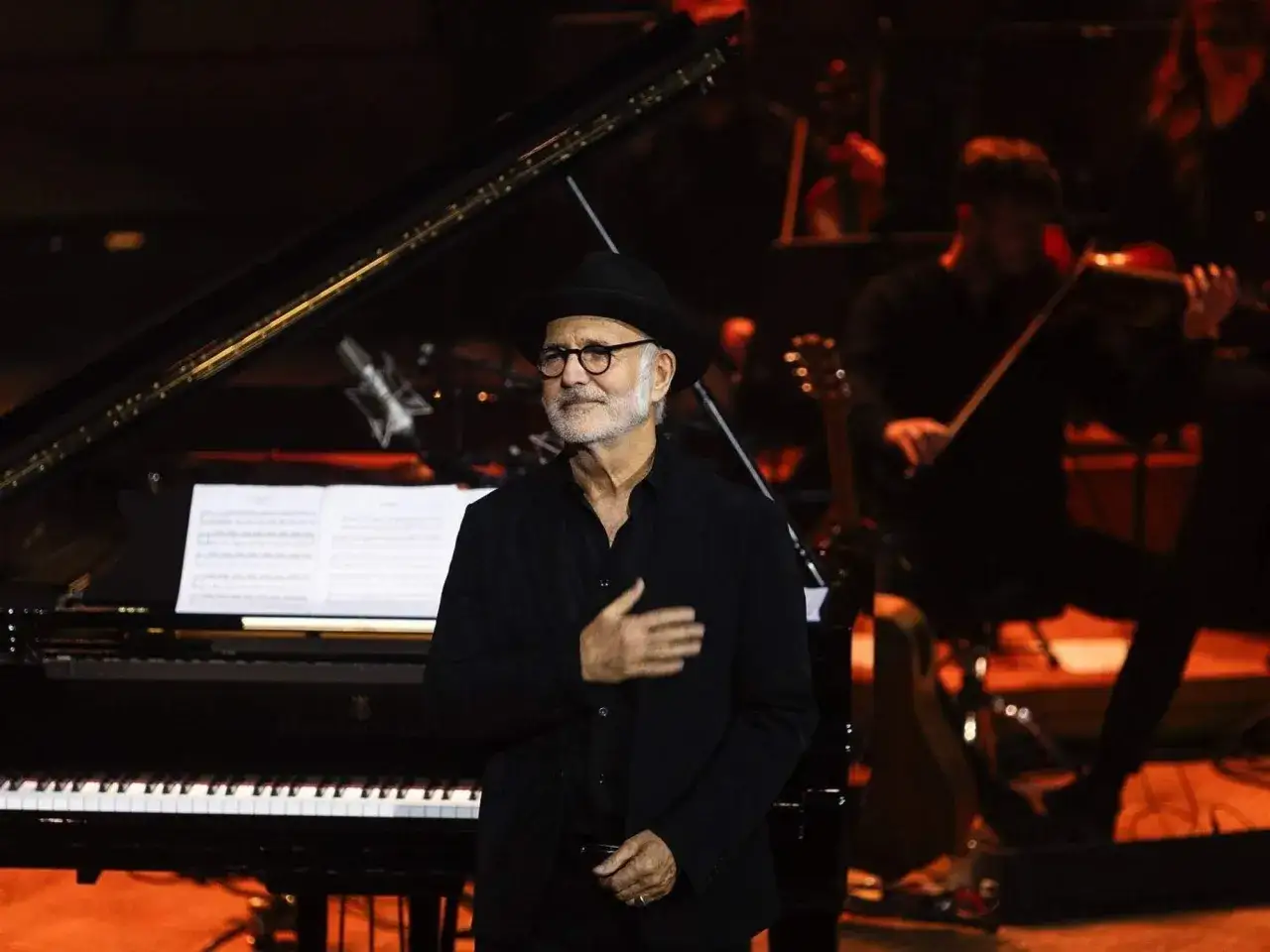 Ludovico Einaudi, con su característica edad y sombrero, dirige una orquesta junto a un piano.