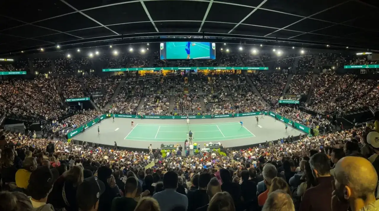 Espectadores animan en un partido de tenis en París. La cancha iluminada y la pantalla gigante muestran la acción del torneo.