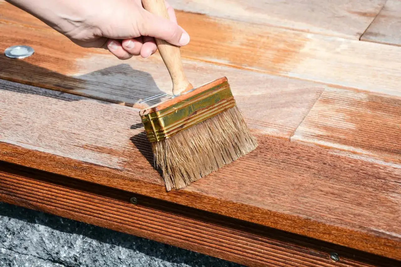 Hand streicht mit breitem Pinsel &uuml;ber ausgeblichenes Holz, um es aufzufrischen. Die Holzoberfl&auml;che wird sichtbar dunkler und erh&auml;lt neuen Glanz.