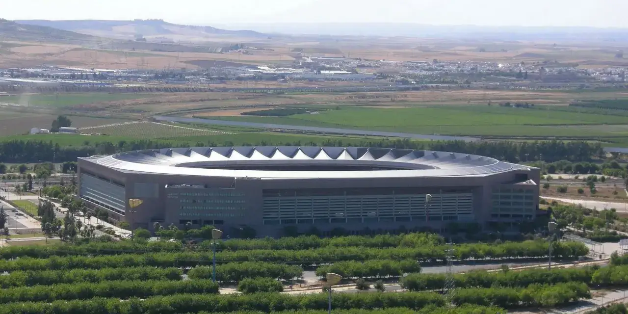 Nowoczesny stadion La Cartuja otoczony zielenią, z widokiem na pola i miasto w oddali.