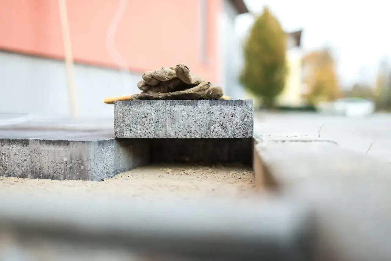 Rękawice robocze leżą na kostce brukowej, czekając na ułożenie. Widać piasek i zarys granicy działki.