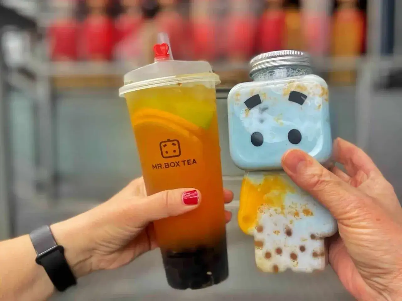 Zwei Hände halten leckere Getränke: ein Bubble Tea von MR.BOX TEA mit Orangenstücken und ein süßer blauer Drink in einer Figurenflasche. Perfekt für einen Tag in Berlin!