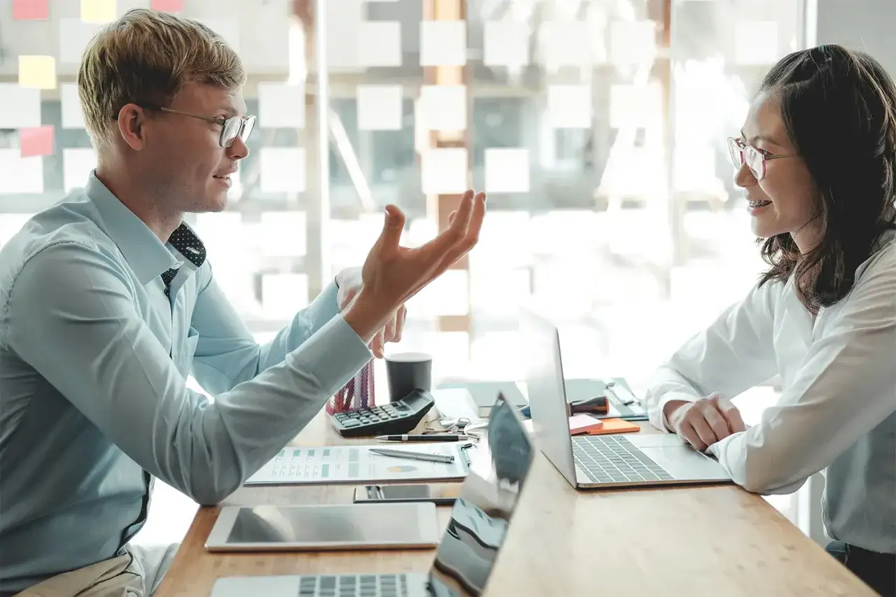 Man gestures while discussing end of year performance review examples with a colleague at a desk with laptops and papers.