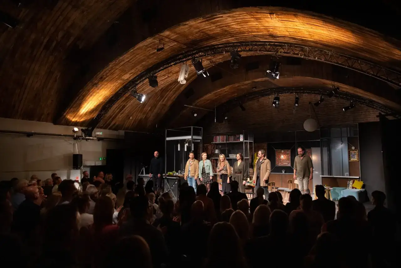 Aktorzy na scenie podczas spektaklu w teatrze. Widownia w ciemnościach, skupiona na przedstawieniu.