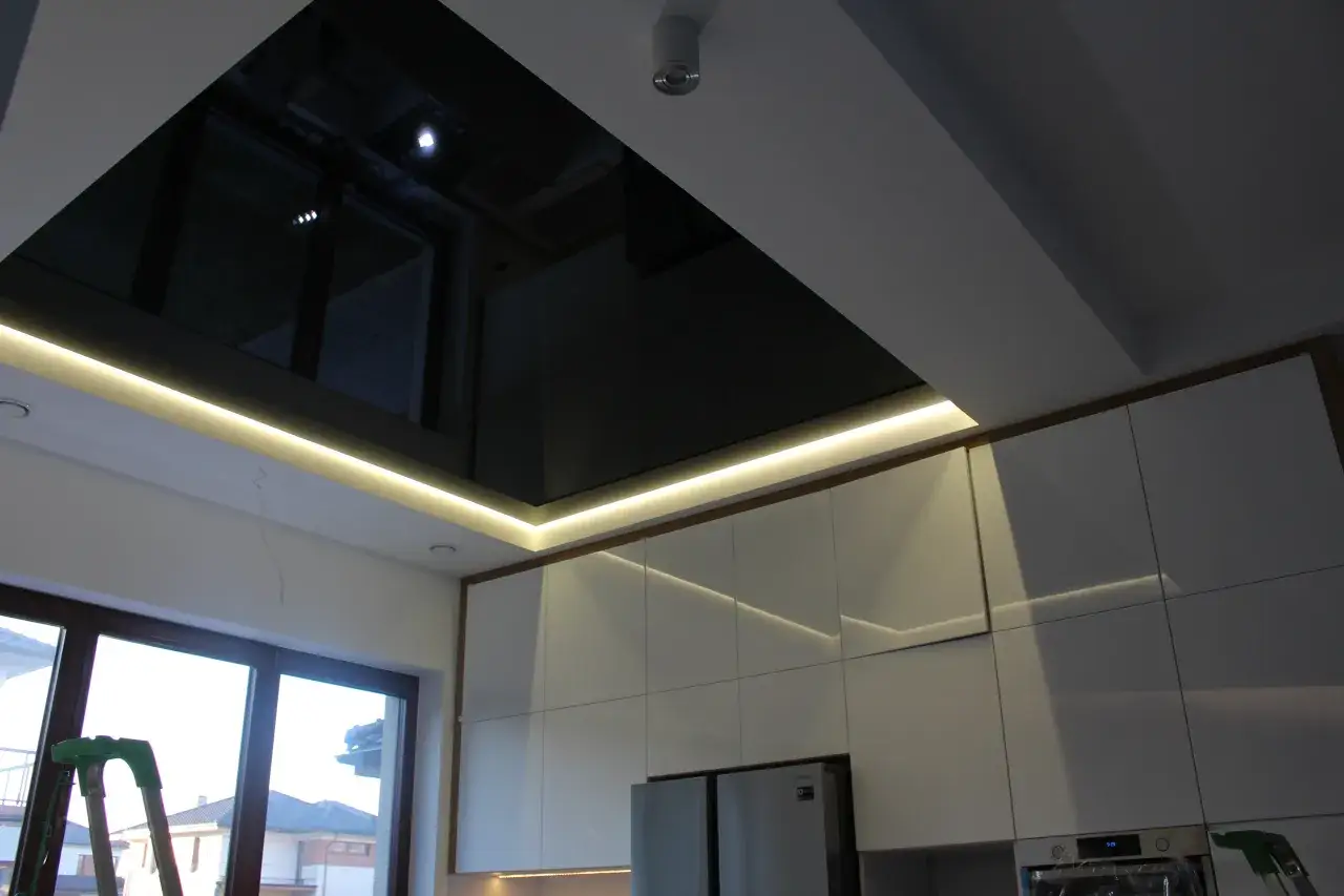 Nowoczesny sufit napinany w kuchni z czarnym połyskiem i ukrytym oświetleniem LED. Białe fronty szafek tworzą elegancką całość.