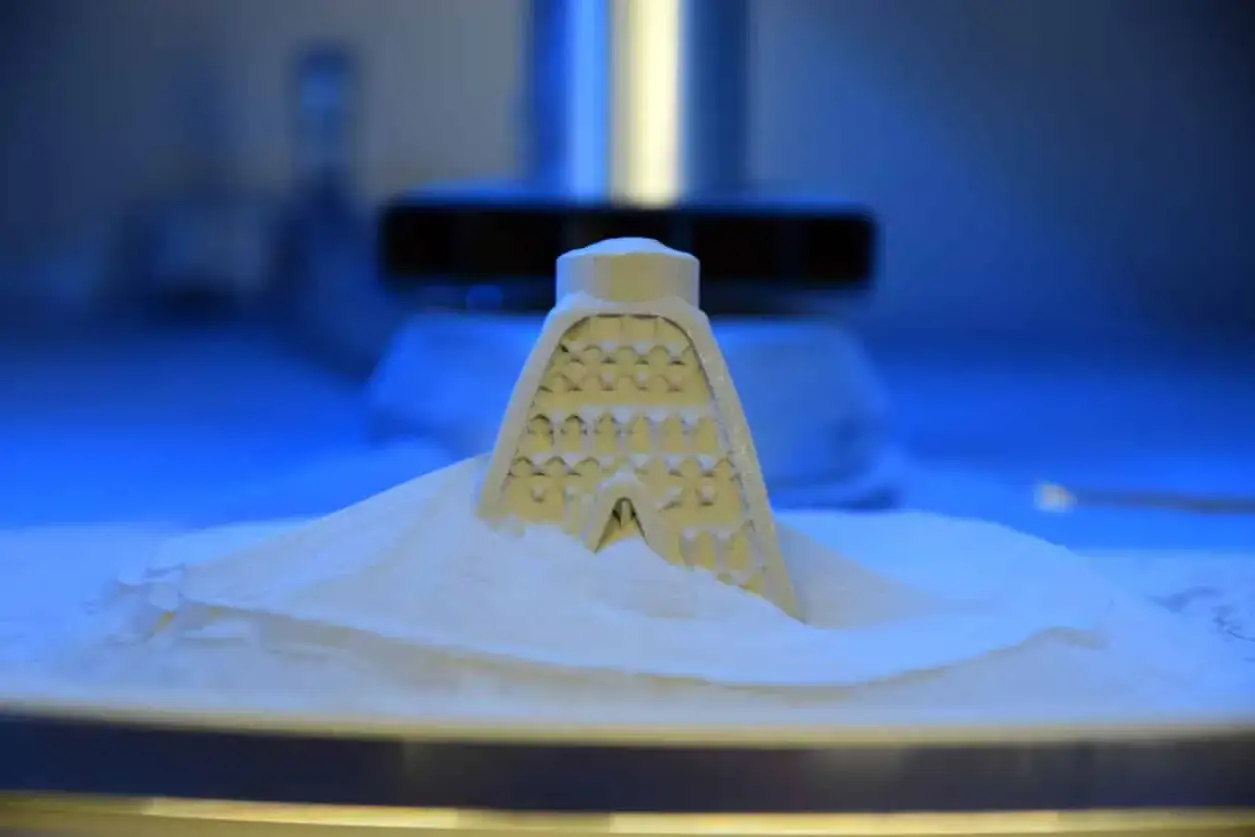 A complex, lattice-like object emerges from a bed of white powder within a laser sintering machine, bathed in cool blue light.