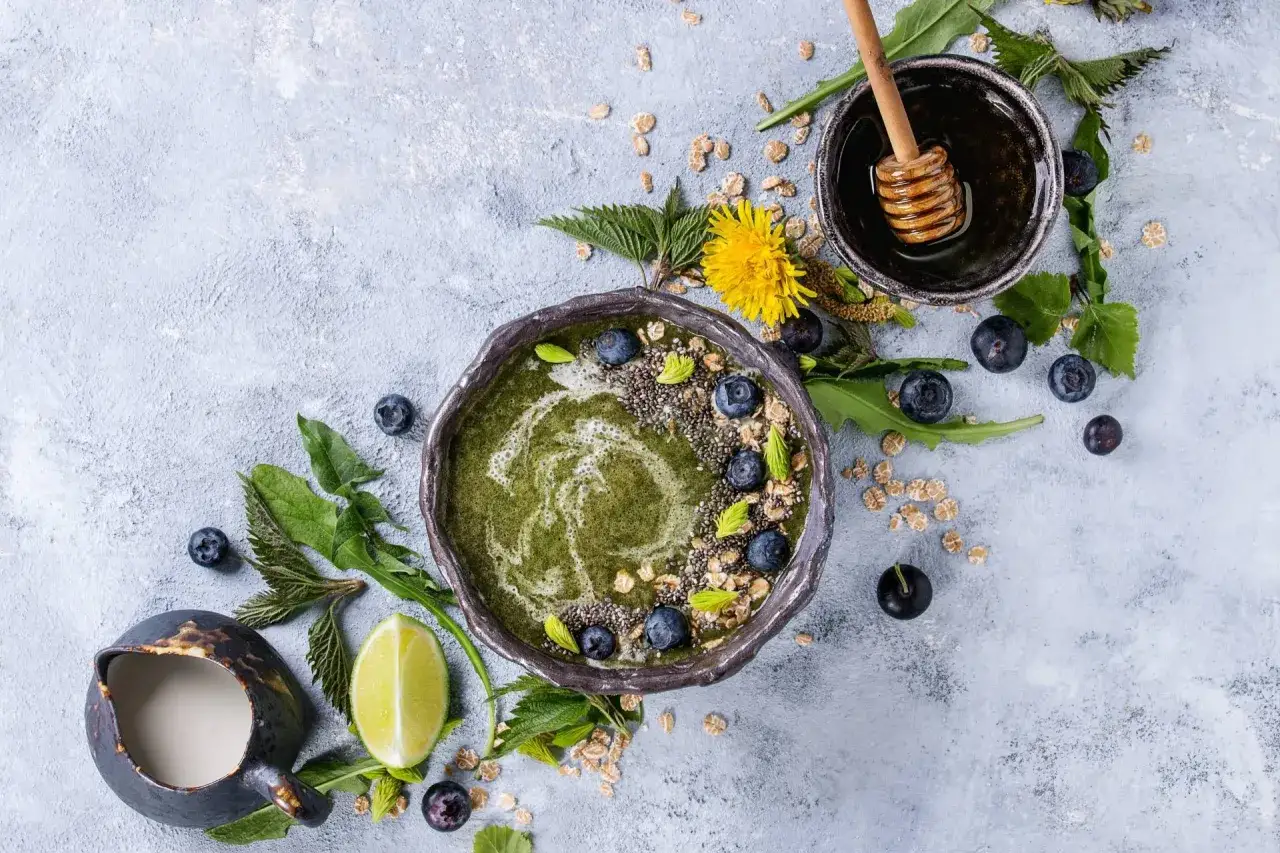 Zielony smoothie bowl z jagodami, nasionami chia i miodem. Wokół liście pokrzywy, podkreślające jej właściwości lecznicze.