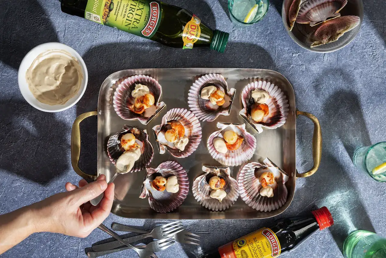 Vieiras a la plancha servidas en sus conchas, con una salsa cremosa. Una mano añade más salsa a una de ellas.