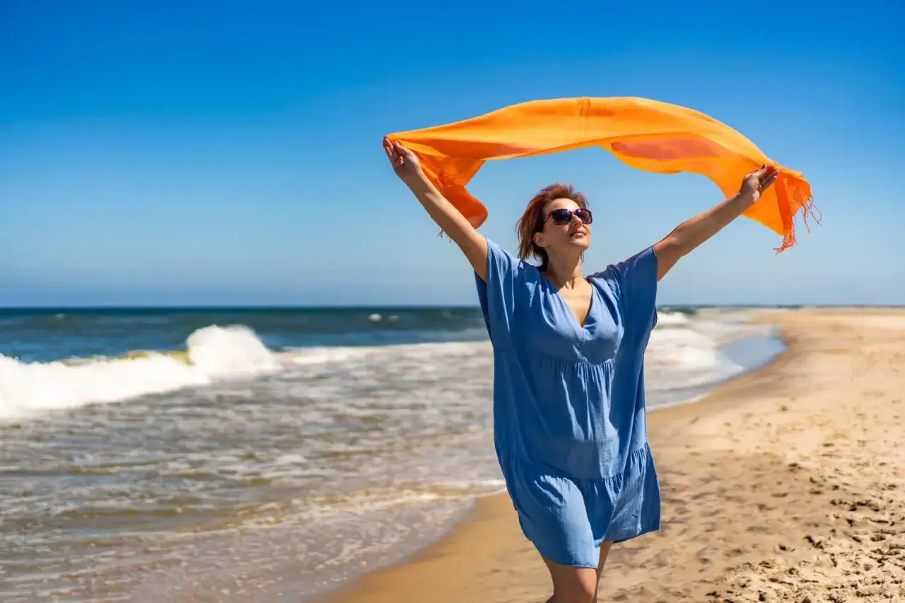 Kobieta w niebieskiej sukience i z pomarańczowym szalem na plaży, ciesząca się świeżym morskim powietrzem. Czy w lesie jest jod? Tu jest jego mnóstwo!