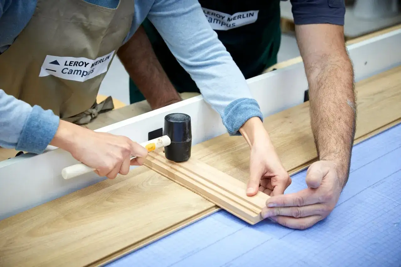 Deux mains assemblent des lames de parquet stratifi&eacute;. L'une utilise un maillet pour ajuster une pi&egrave;ce, l'autre guide.