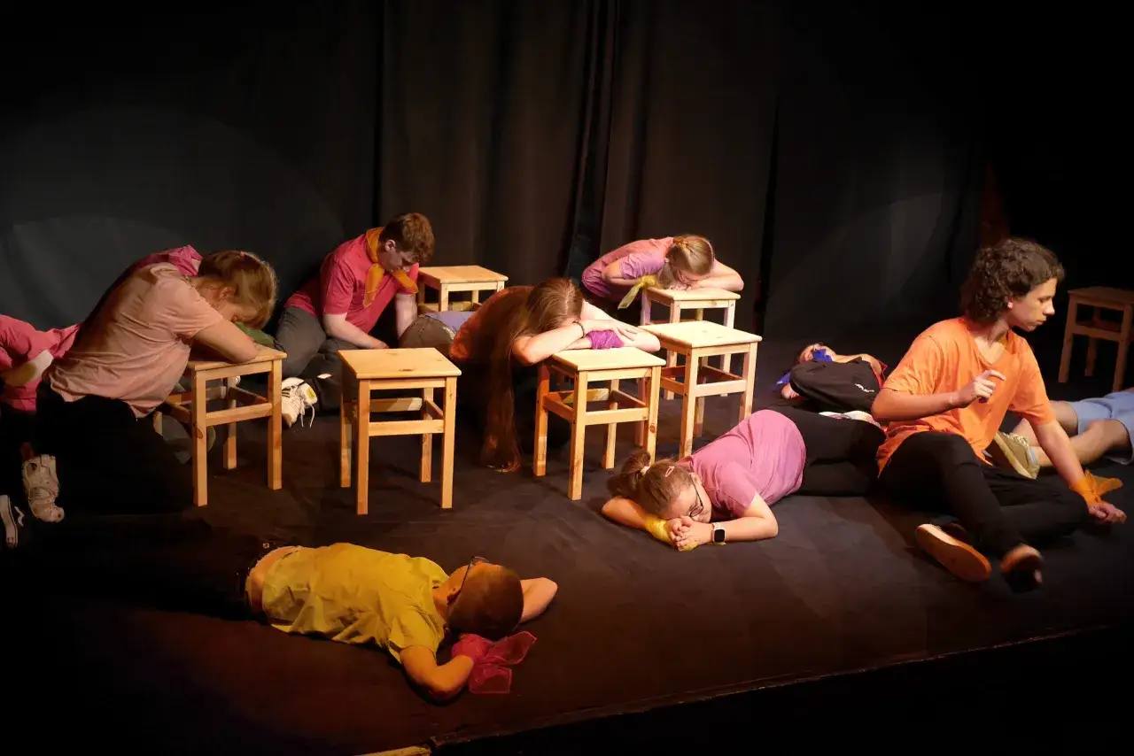 Dzieci na scenie, w ramach przedstawienia teatralnego, leżą lub pochylają się nad drewnianymi stołkami, tworząc scenę pełną emocji.