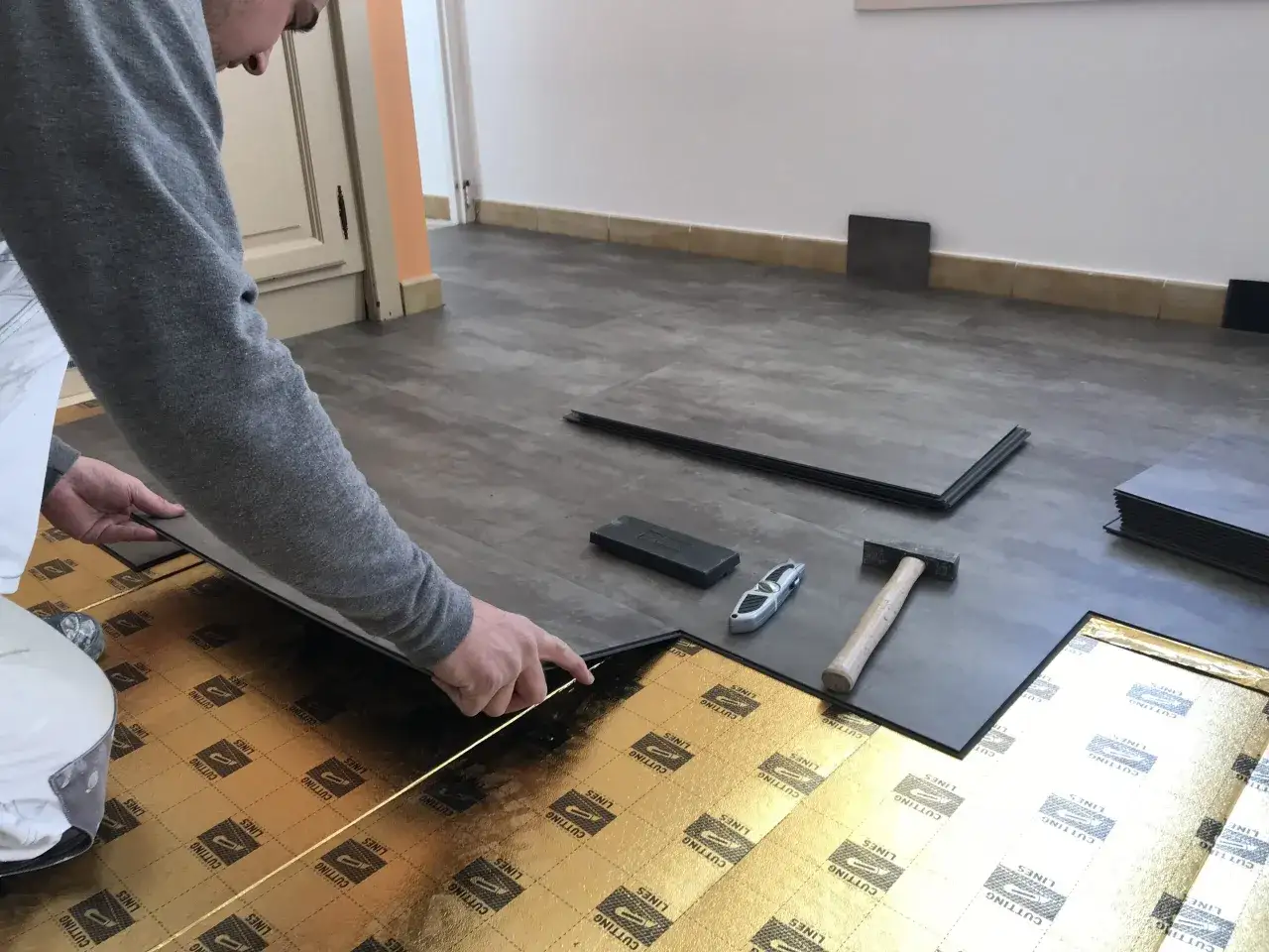 Pose d'une lame PVC clipsable sur une sous-couche dor&eacute;e. Un marteau et un cutter sont &agrave; proximit&eacute;.