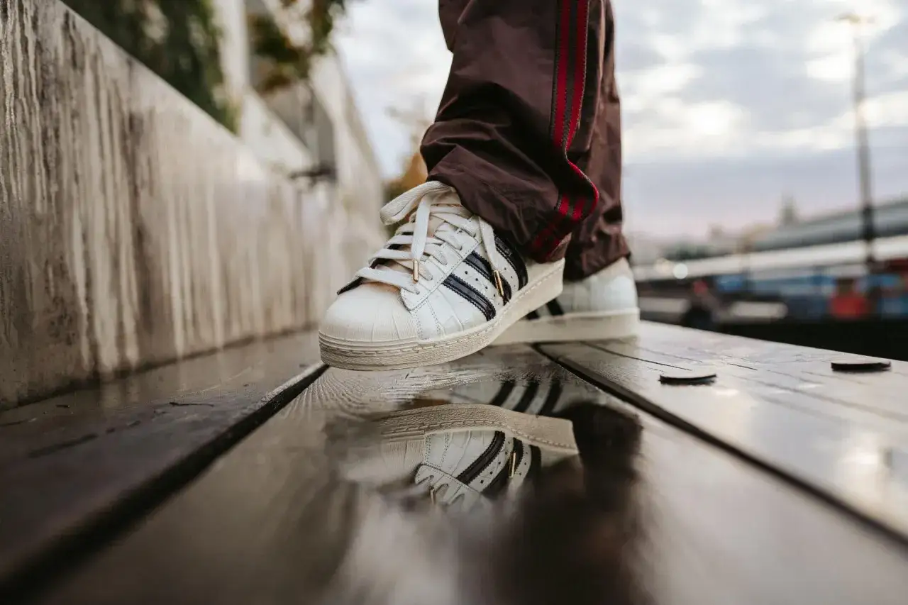 Wei&szlig;e Adidas Superstar Sneaker mit Kroko-Pr&auml;gung, Teil einer neuen Kollektion, spiegeln sich im nassen Holz.