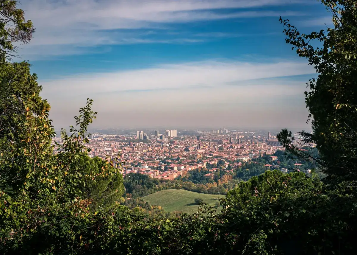 Vista panoramica dei colli bolognesi con la città in lontananza, incorniciata da alberi verdi e un cielo azzurro con nuvole.