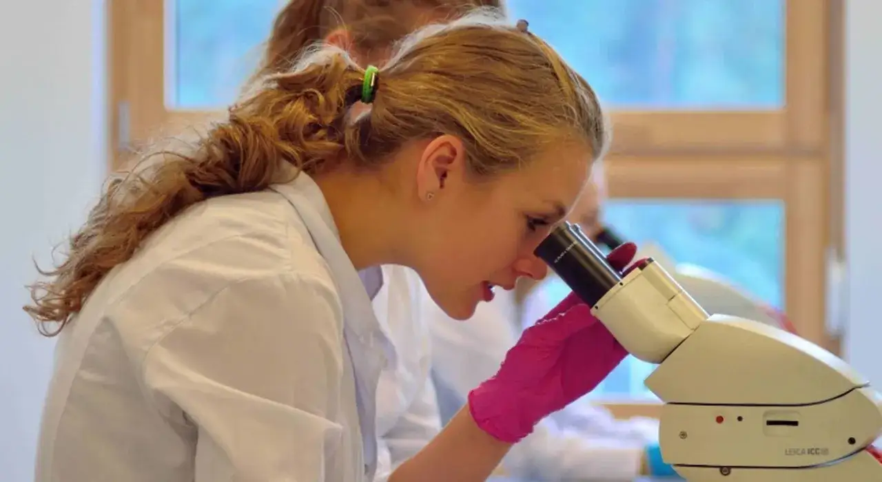 Dziewczyna w różowej rękawiczce pochyla się nad mikroskopem, zgłębiając tajniki biologii podczas studiów.