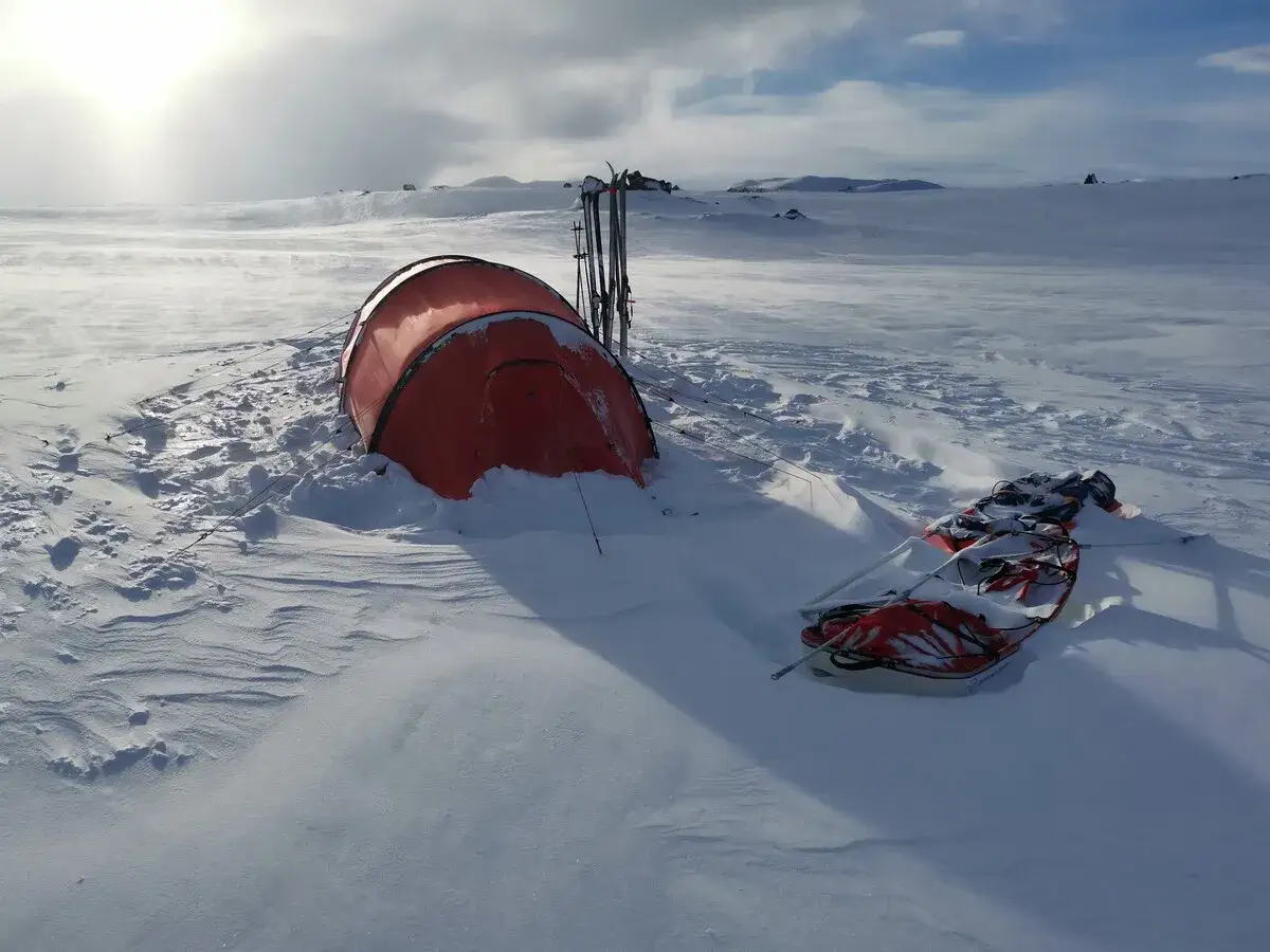 Ein rotes Wintercamping Zelt steht im Schnee, daneben Skier und ein Schlitten. Die Landschaft ist weit und verschneit.