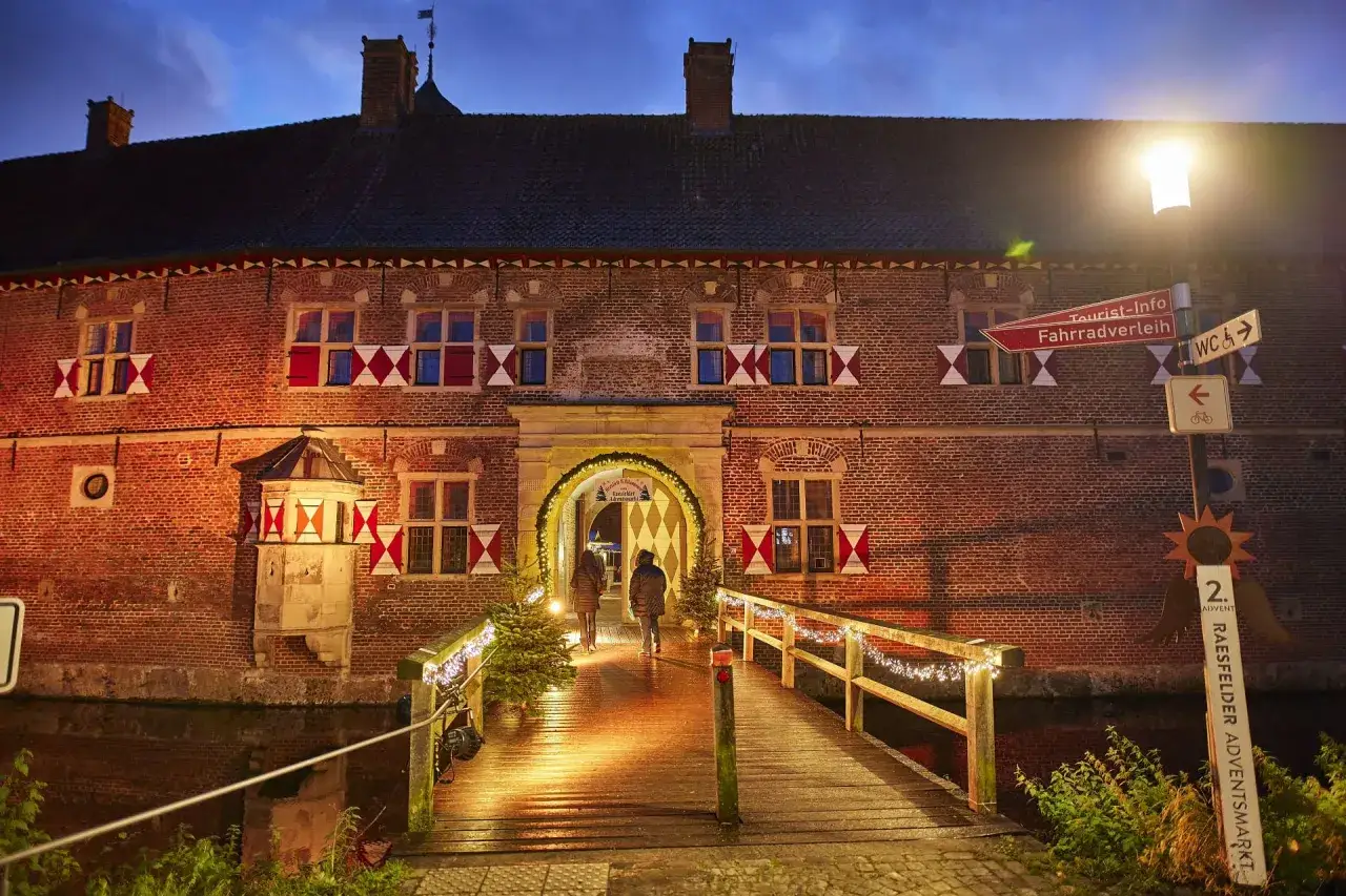 Ein beleuchteter Holzsteg führt zum Wasserschloss, wo der Raesfelder Adventsmarkt mit Lichtern und Weihnachtsbäumen lockt.