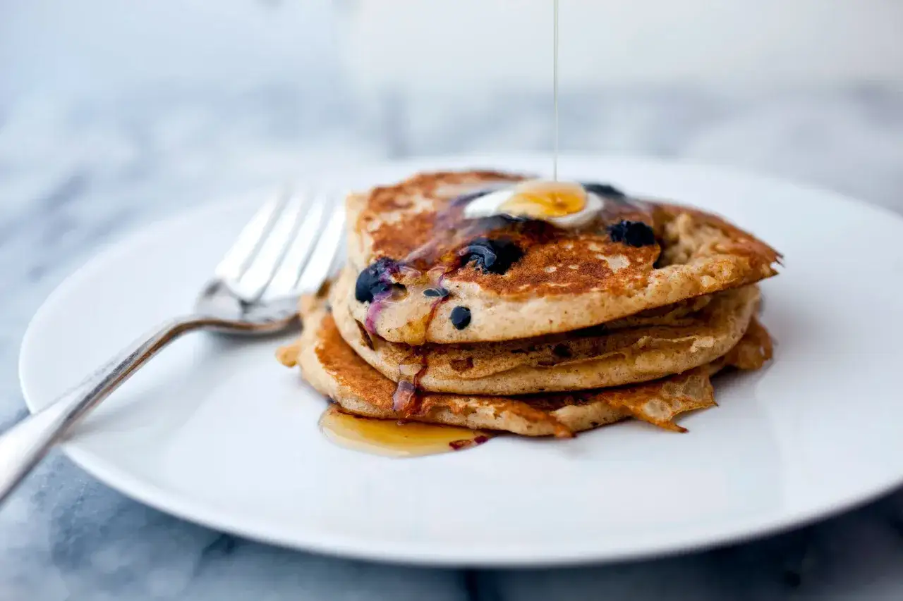 Ein Stapel gesunde Pancakes mit Blaubeeren, betr&auml;ufelt mit Sirup und einem Klecks Butter. Daneben liegt eine Gabel.