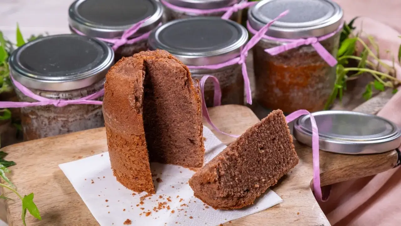 Ein Anschnitt eines Schokoladenkuchens liegt auf einem Holzbrett, daneben ein St&uuml;ck. Im Hintergrund stehen mehrere Gl&auml;ser mit Kuchen im Glas, verziert mit rosa B&auml;ndern.