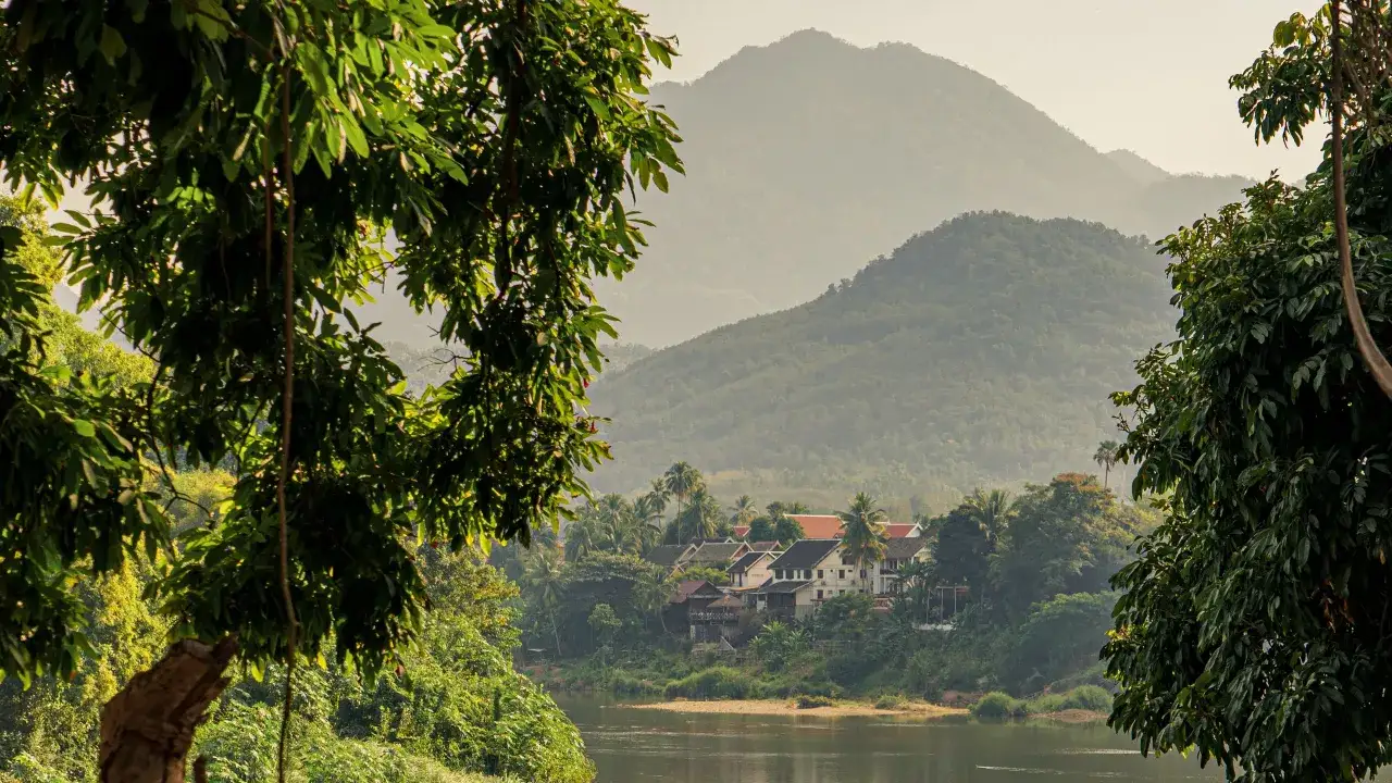 Grüne Hügel und ein Flussufer mit Häusern und Palmen, ein idyllischer Ort für Laos Backpacking.