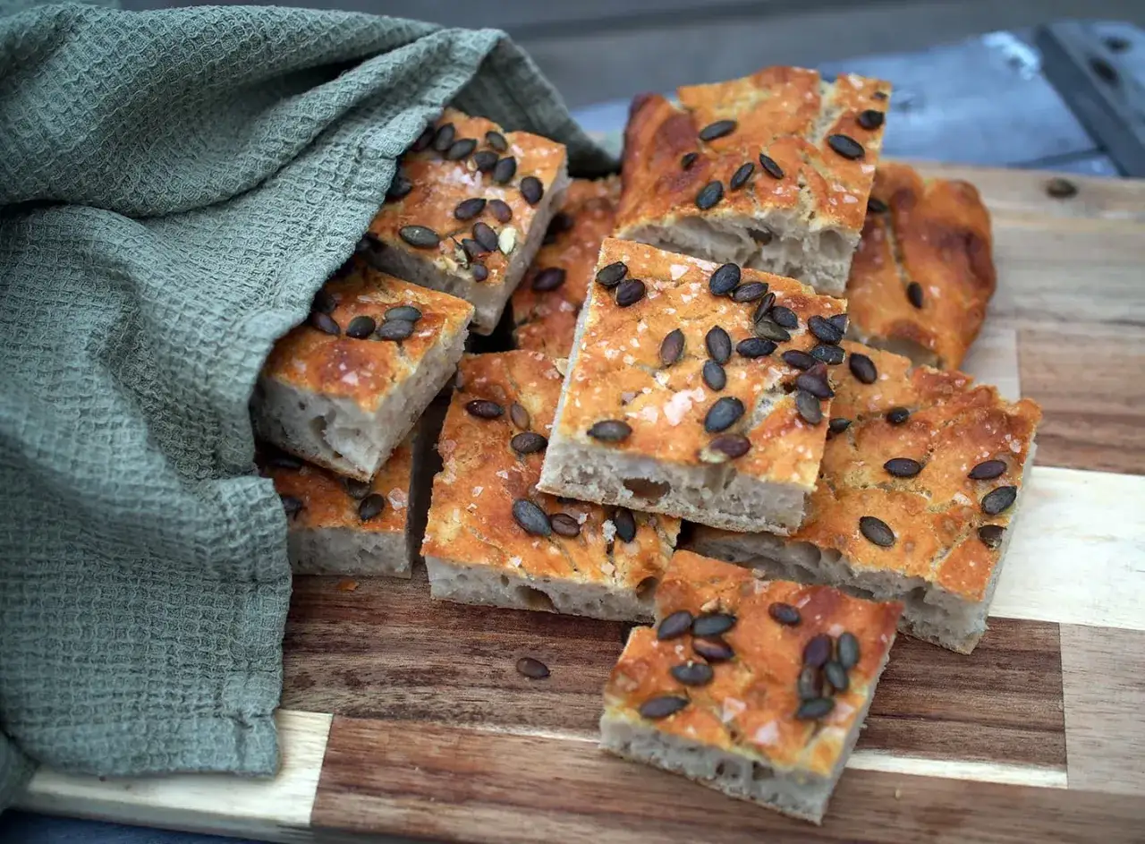 Focaccia med pumpakärnor och havssalt, bakad med dinkelmjöl istället för vetemjöl. Mjukt inlindad i en grön handduk.