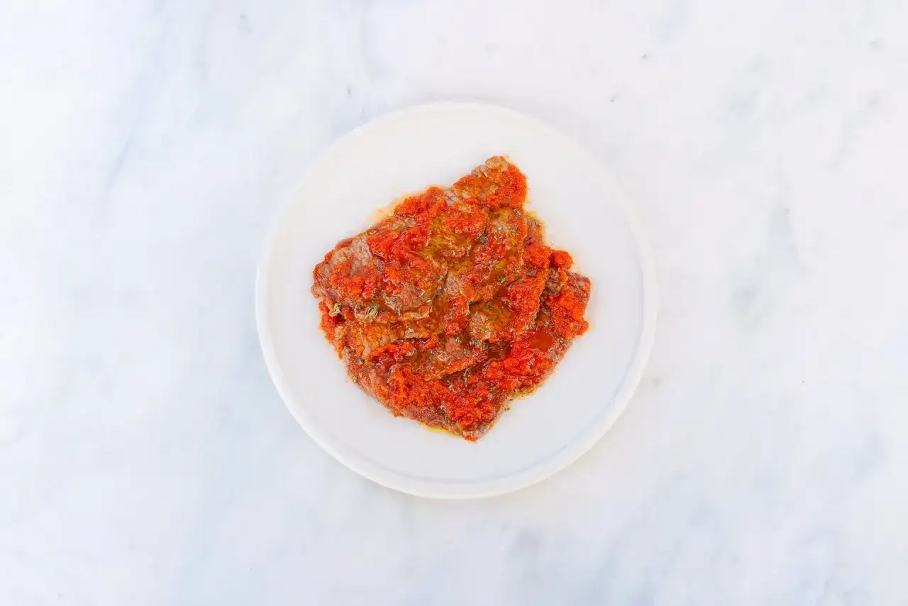 Ein d&uuml;nnes St&uuml;ck Fleisch, bedeckt mit einer leuchtend roten Tomatenso&szlig;e und Kr&auml;utern, serviert auf einem wei&szlig;en Teller. Ein Hauch von pizzaiola original italienisch.