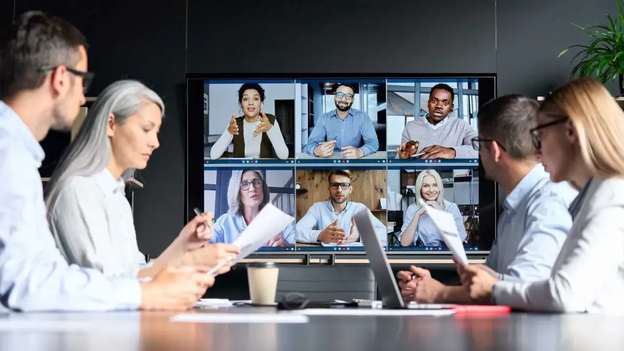 A diverse team collaborates remotely, showcasing adaptability in leadership. Six faces appear on a screen, while three colleagues in the foreground engage in discussion.