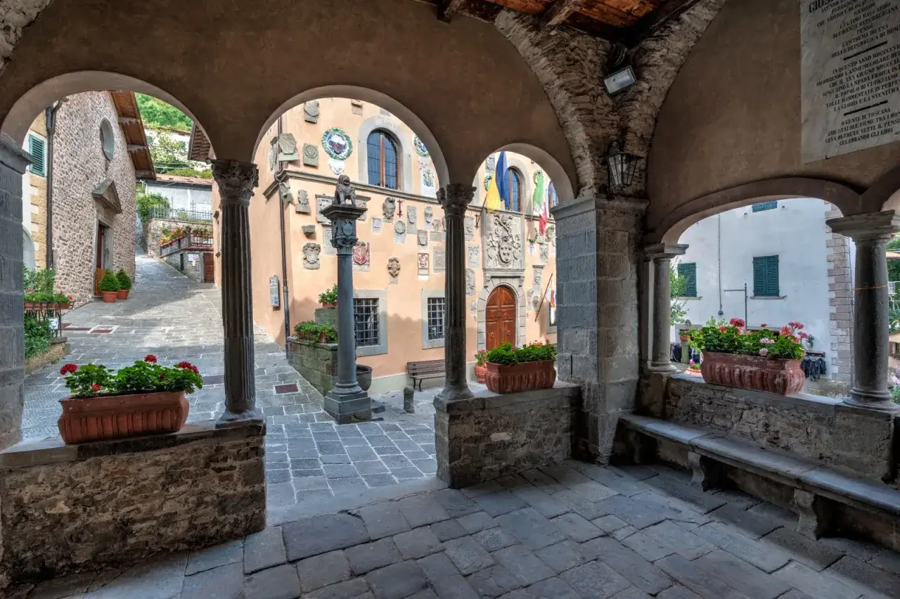 Un vicolo acciottolato tra antichi borghi vicino Abetone, con case decorate da stemmi e fiori rossi che sbocciano in vasi di terracotta.