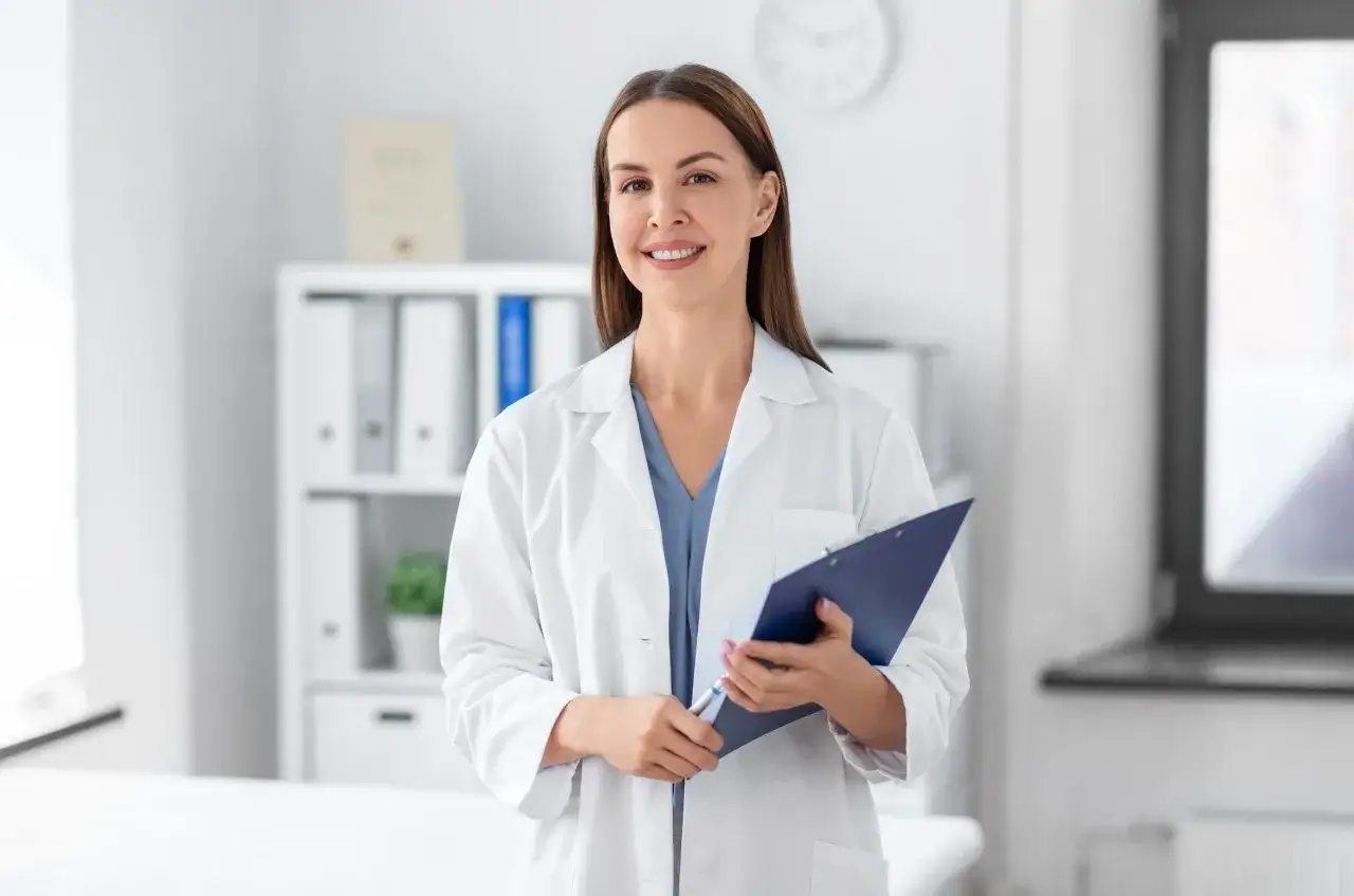 Uśmiechnięta lekarka w białym kitlu trzyma teczkę. Czy do poradni dermatologicznej potrzebne jest skierowanie? Zapytaj specjalistę.