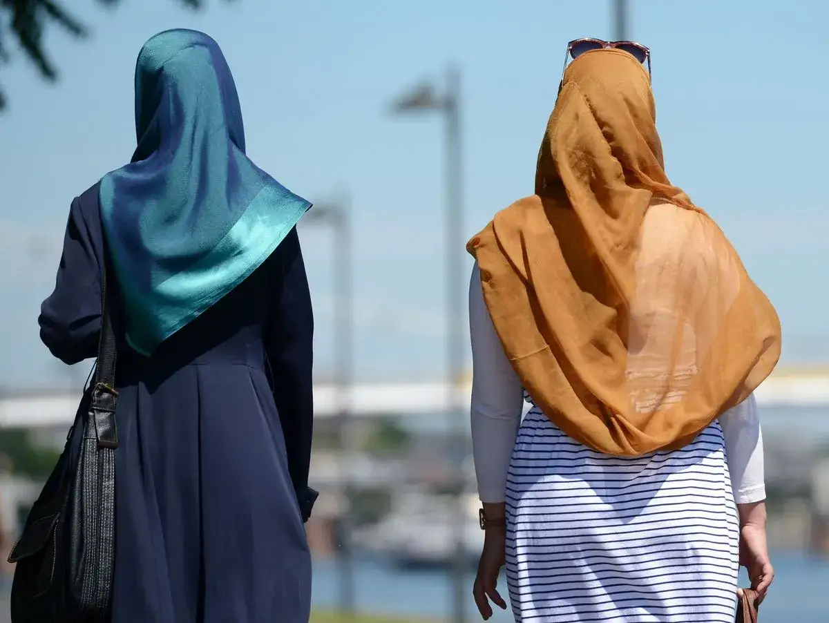 Zwei Frauen mit Kopftuch gehen nebeneinander. Eine tr&auml;gt ein blaues Kleid, die andere ein gestreiftes Kleid.