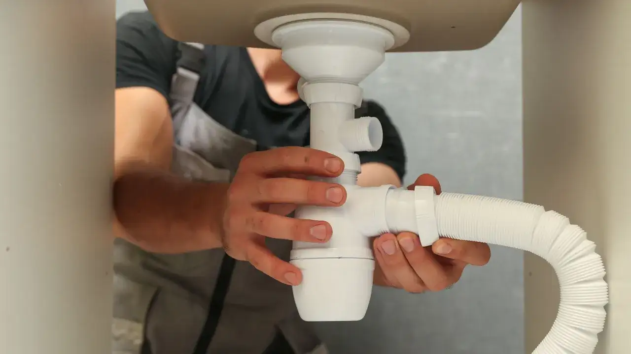 Sch&eacute;ma montage siphon lavabo : un plombier assemble les pi&egrave;ces d'un siphon blanc sous un &eacute;vier.