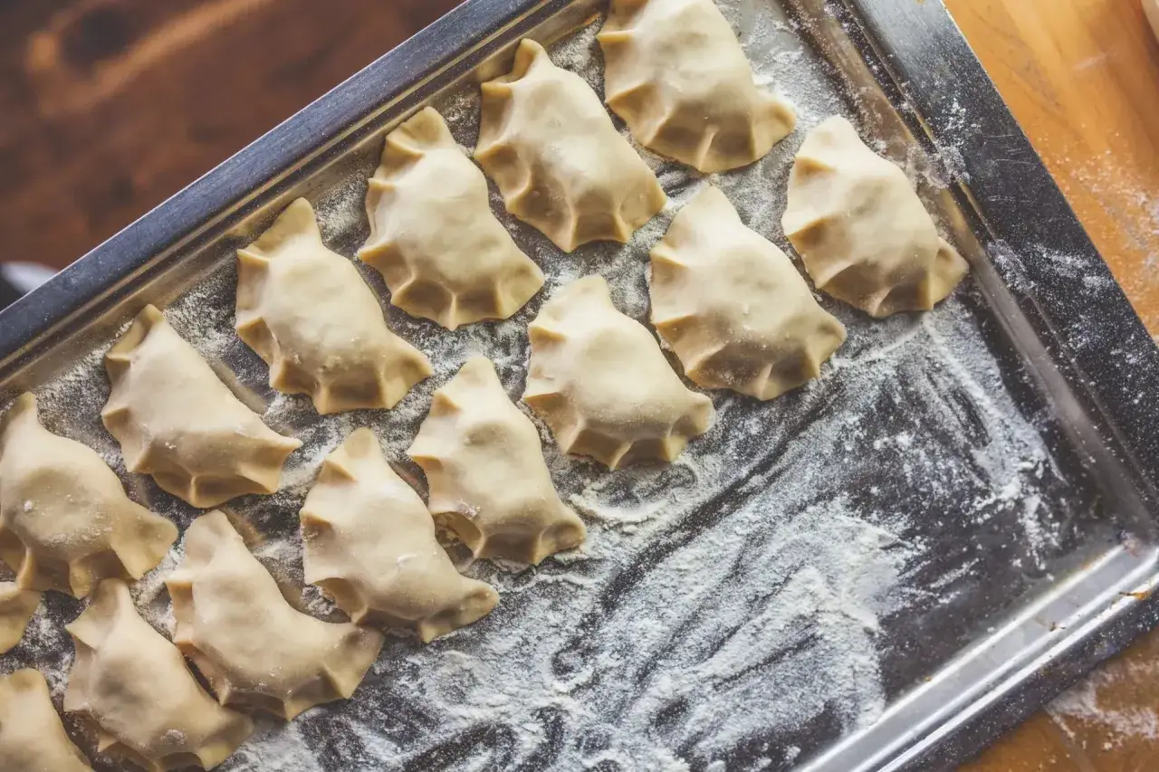 Gotowe do gotowania pierogi z serem leżą na metalowej tacy, posypanej mąką.