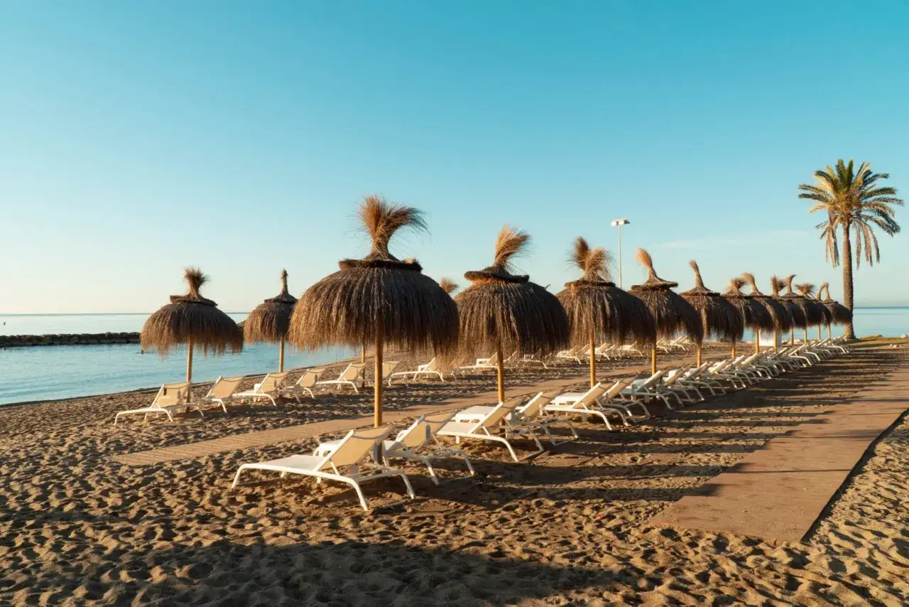 Plaża w Marbelli z rzędem leżaków i parasoli z trawy. Słońce oświetla piasek i turkusowe morze.
