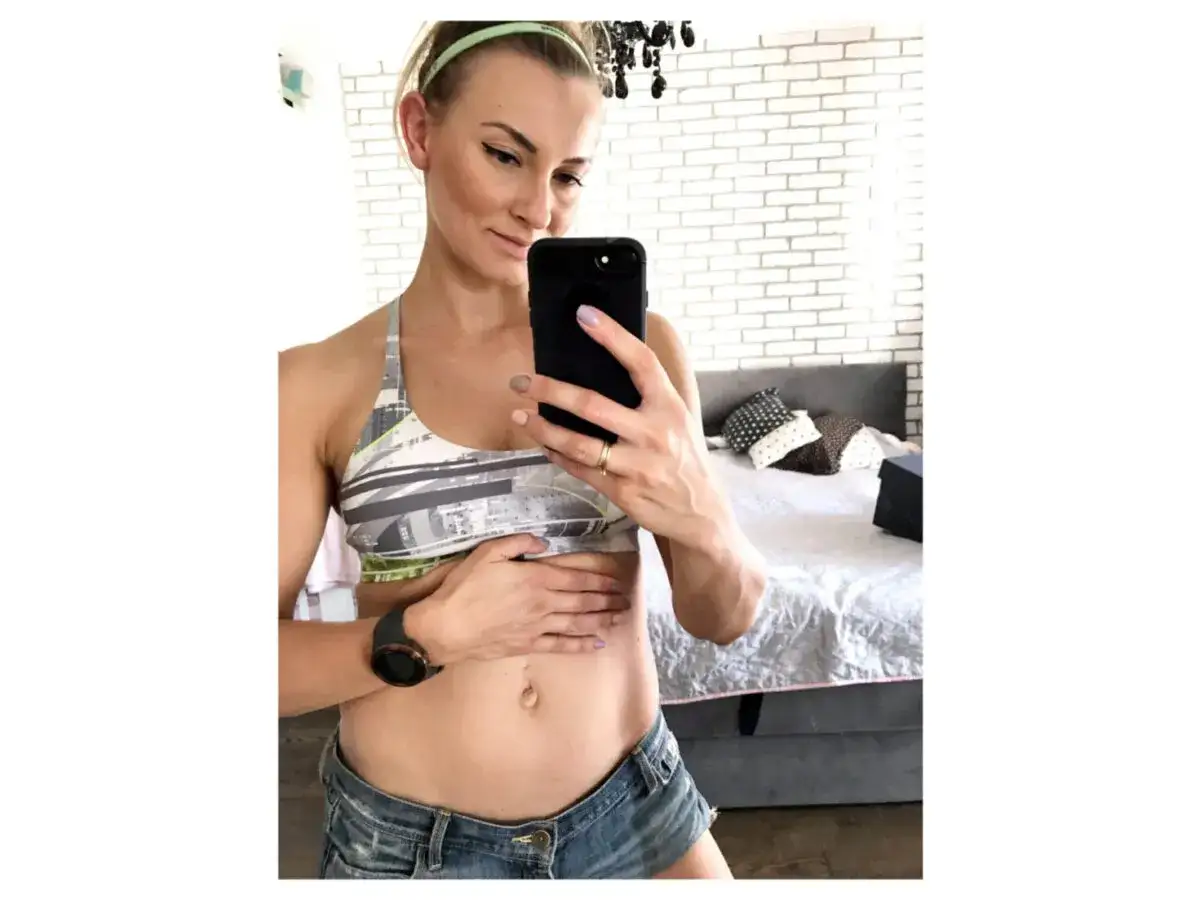 Kobieta w sportowym topie i kr&oacute;tkich spodenkach robi selfie, pokazując brzuch. Może to być część jej ćwiczeń na przepuklinę pępkową.