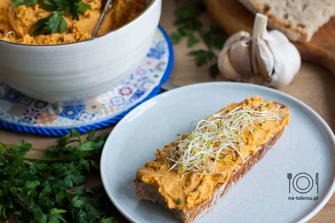 Pyszna pasta z kurczaka na kromce chleba, posypana kiełkami. Obok miska z pastą, czosnek i natka pietruszki.