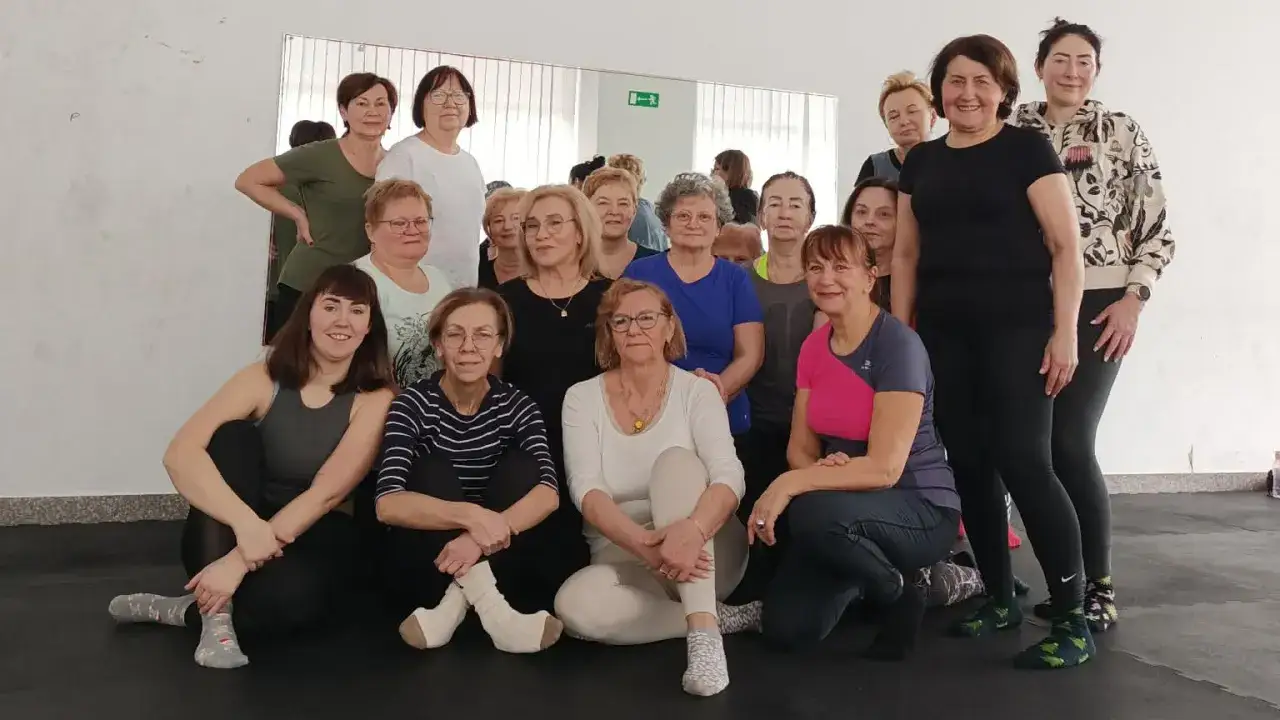 Grupa uśmiechniętych kobiet, uczestniczek zajęć dla seniorów, pozuje do zdjęcia po treningu.