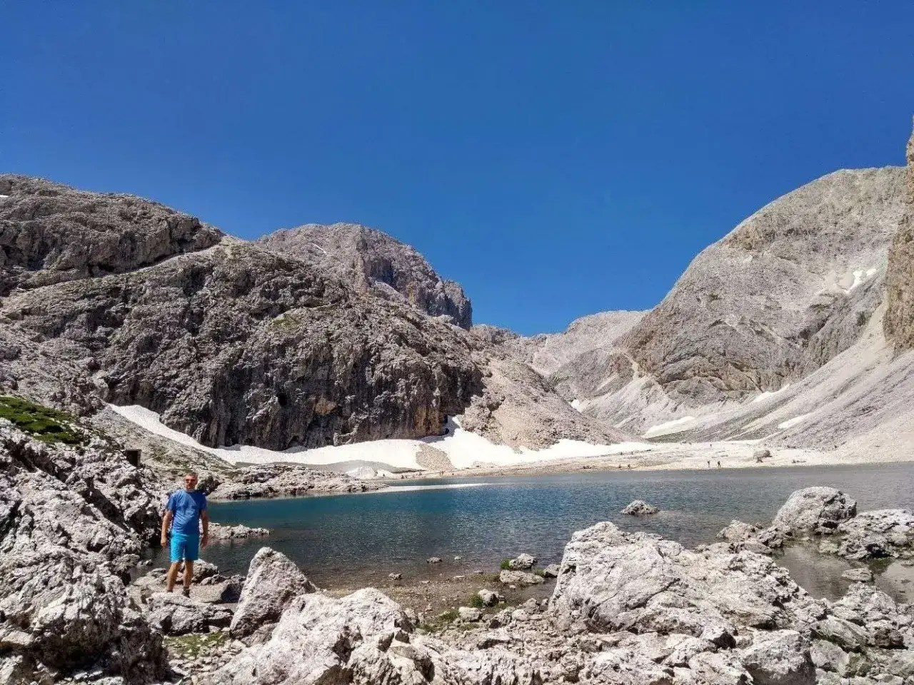 Un uomo in piedi su rocce bianche ammira il **lago di Antermoia**, circondato da montagne imponenti e chiazze di neve.