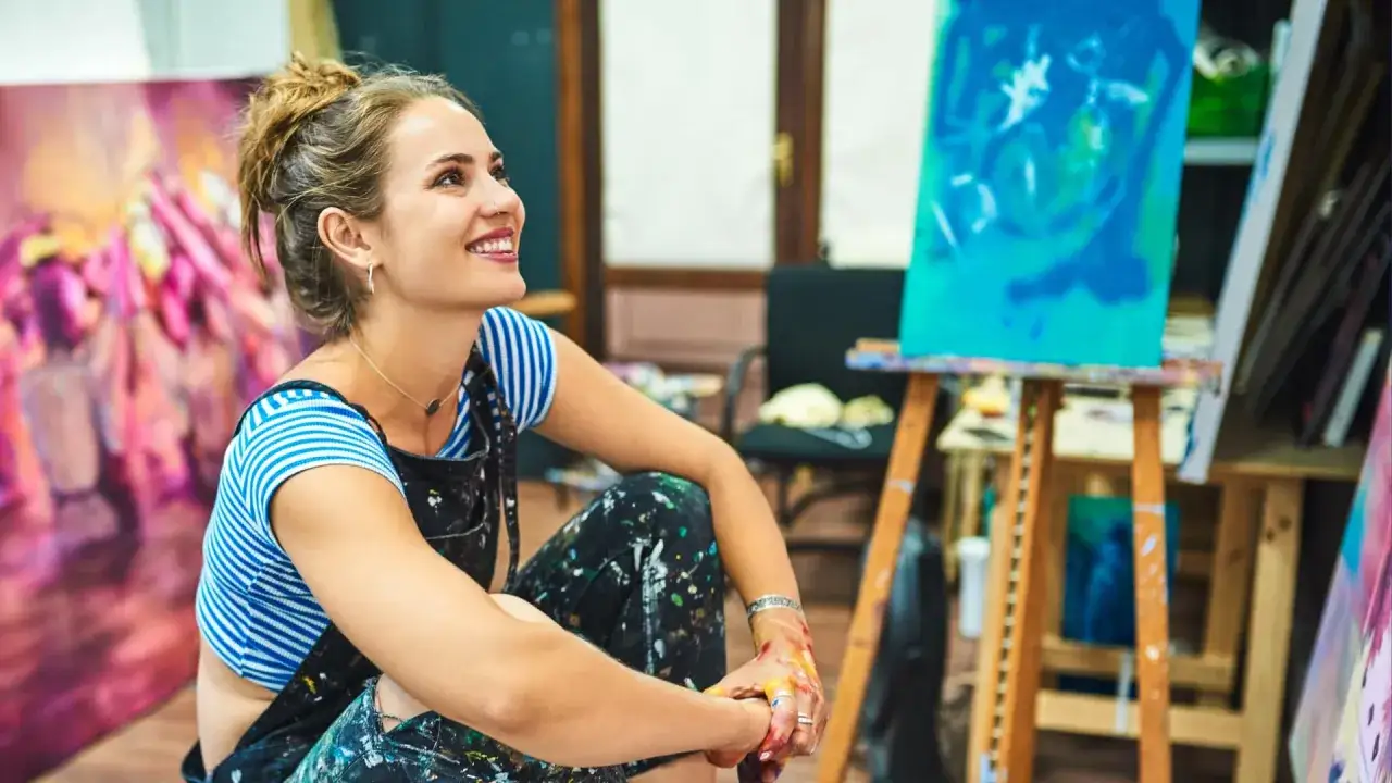 Une artiste entrepreneur souriante, assise au milieu de ses toiles colorées, son tablier maculé de peinture.