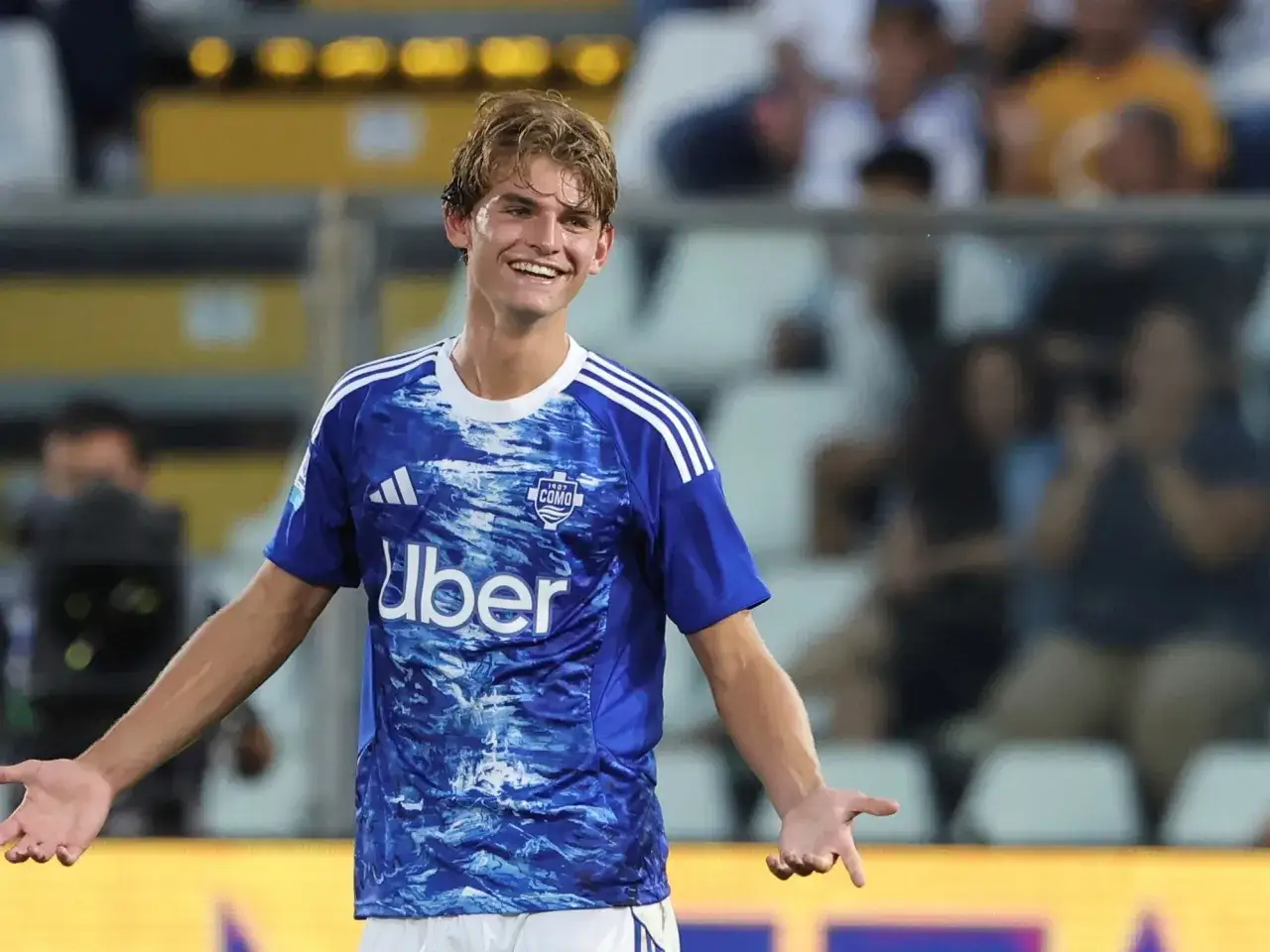 Joven jugador de f&uacute;tbol, Nico Paz, sonr&iacute;e con los brazos extendidos en un partido.